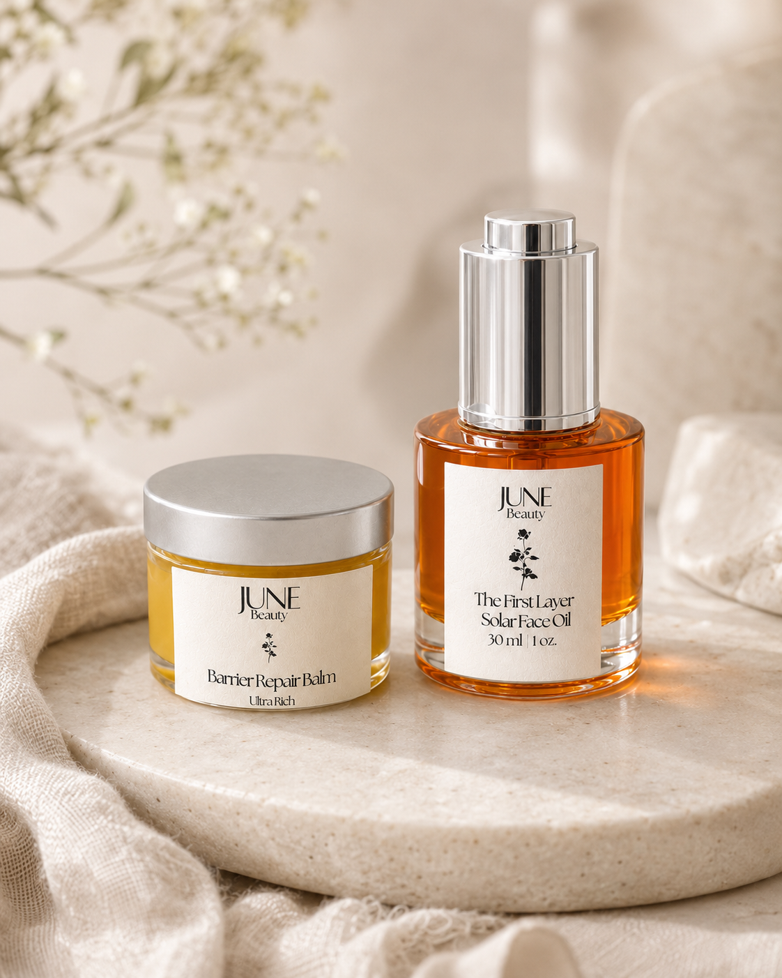 Two June Beauty skincare products, Barrier Repair Balm and The First Layer Solarise Oil, displayed on a round stone slab with draped beige fabric and soft floral elements in the background.